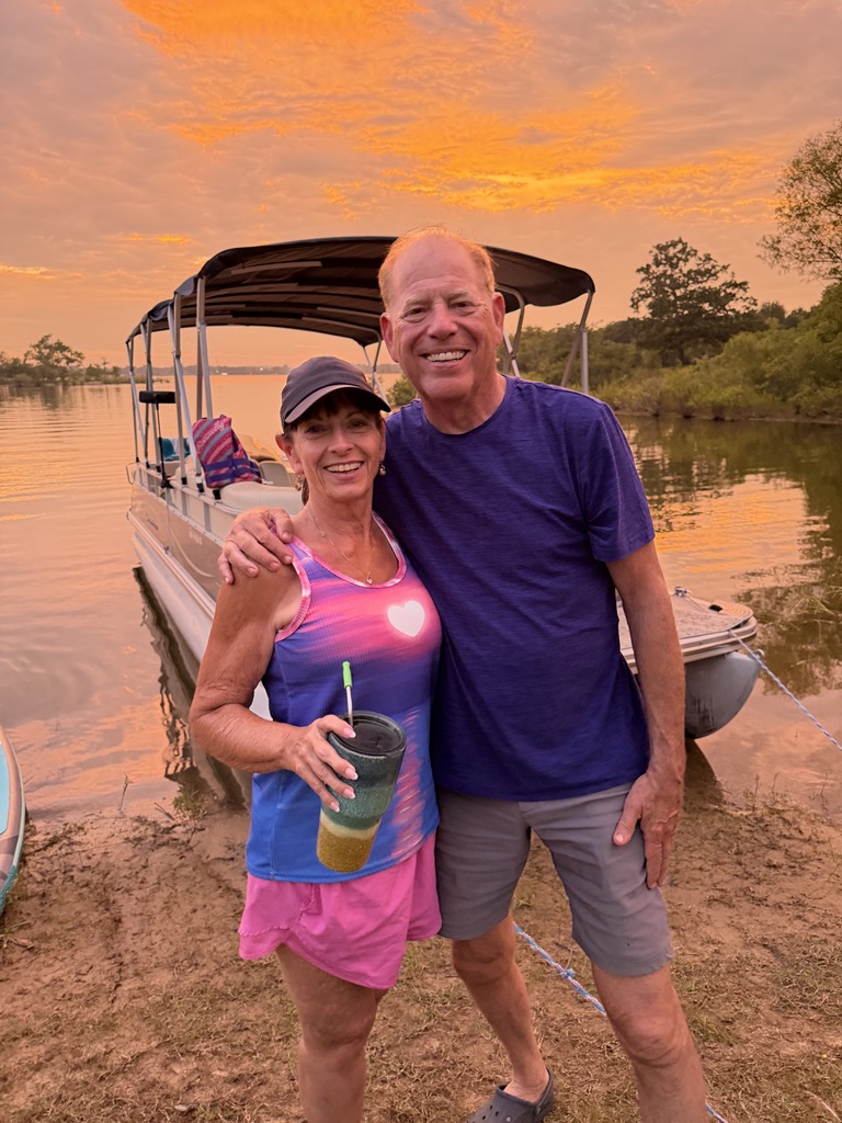 Tom and Deb smiling beside their pontoon boat at the lake under an orange sunset